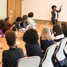 Eine Gruppe von Büroangestellten in einem Konferenzraum, die einer Frau am Whiteboard zuhören